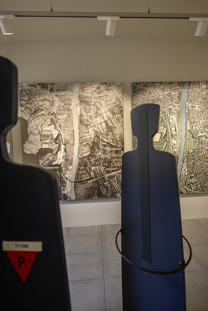 Eine Aufnahme des Ausstellungsbereichs KZ-Komplex der Gedenkstätte Neckarelz. Zu sehen sind ausgeschnittene Schwarze Silhoutten von Personen vor Luftaufnahmen des KZ-Komplexes im Vergleich damals zu heute.